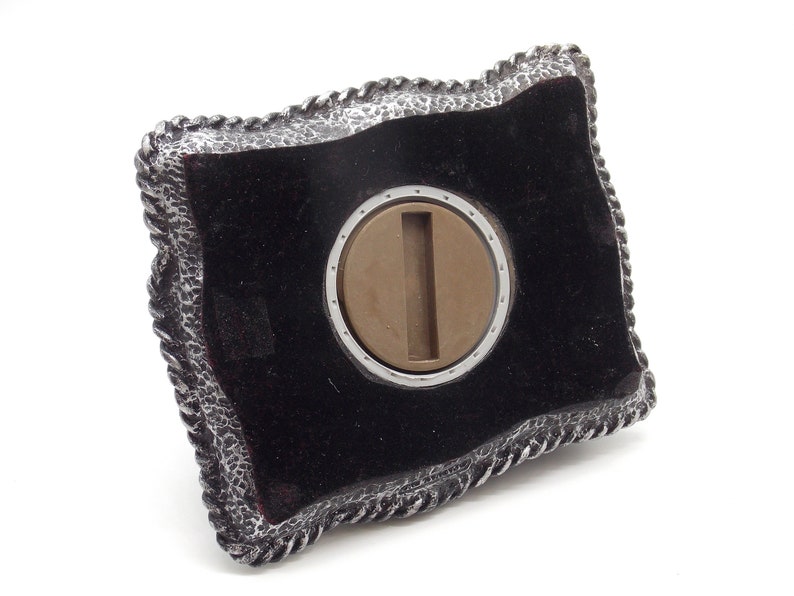 May include: A silver belt buckle with a black velvet inlay and a silver-toned metal insert. The buckle has a rope-like design around the edge.