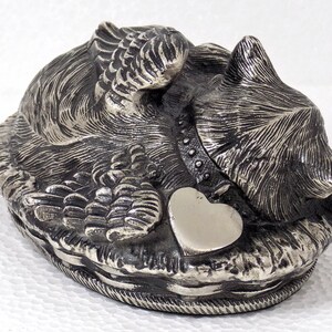 K&ouml;nnte beinhalten: Silberne Metallfigur eines Hundes mit Fl&uuml;geln, der mit einem herzf&ouml;rmigen Anh&auml;nger liegt. Der Hund ist detailliert mit Fell und hat ein Halsband.