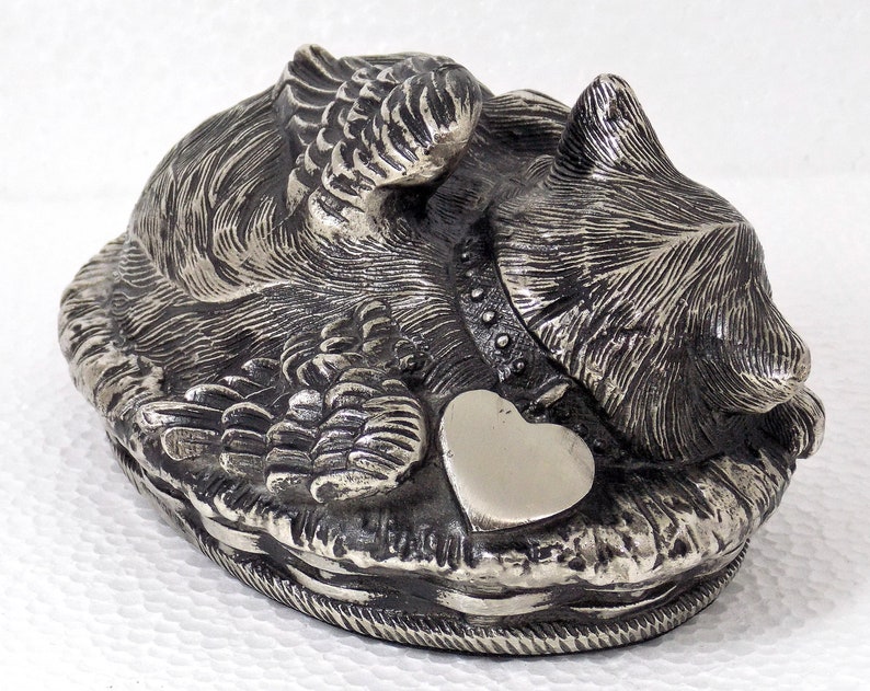 K&ouml;nnte beinhalten: Silberne Metallfigur eines Hundes mit Fl&uuml;geln, der mit einem herzf&ouml;rmigen Anh&auml;nger liegt. Der Hund ist detailliert mit Fell und hat ein Halsband.