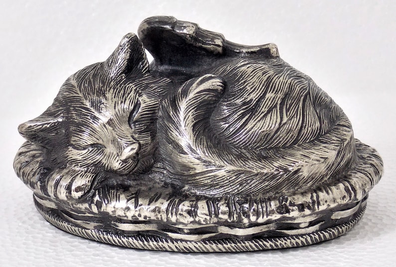 K&ouml;nnte beinhalten: Eine silberfarbene Metallfigur eines schlafenden Katers auf einem Bett. Der Kater ist zusammengerollt, mit dem Kopf auf seinen Pfoten ruhend. Das Bett ist mit einem gewebten Muster verziert.