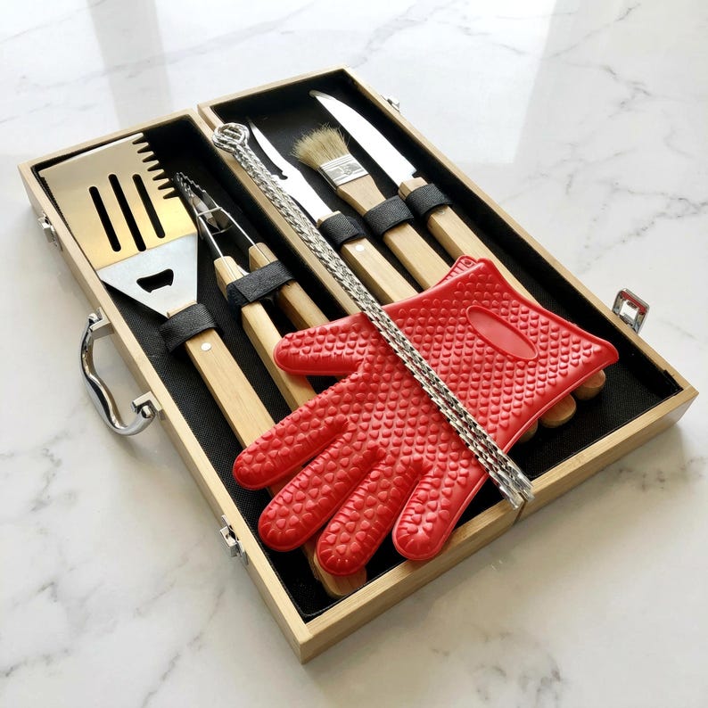 May include: A wooden barbecue tool set with stainless steel utensils, a red silicone glove, and a metal chain. The set includes a spatula, tongs, a brush, and knives. The tools have wooden handles and are secured in a wooden box.