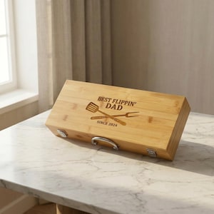 May include: A rectangular bamboo grilling tool box with the text "BEST FLIPPIN' DAD SINCE 2024" and crossed spatula and fork design. The box has a silver handle and latches.