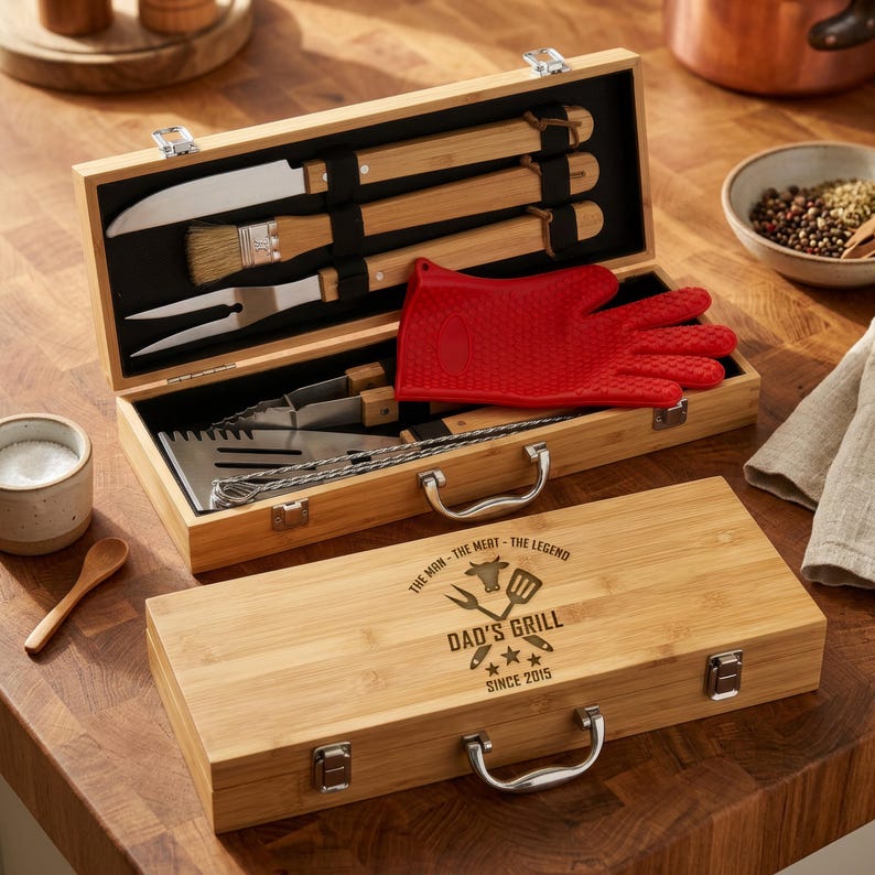 May include: A bamboo barbecue tool set in an open wooden case, featuring a knife, brush, fork, spatula, and tongs. A red silicone glove rests inside. The closed case has the text "DAD'S GRILL" and "SINCE 2015".