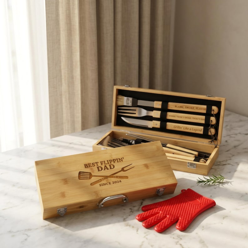 May include: A bamboo barbecue tool set with the text "Best Flippin' Dad Since 2024" engraved on the lid. The set includes a spatula, fork, brush, knife, and tongs. A red silicone oven mitt is also present.