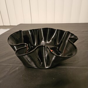 May include: A handcrafted bowl made from a recycled vinyl record. The bowl is black with a glossy finish and a wavy, ruffled edge. The center of the record is visible, with red and orange label details. A unique decorative piece.