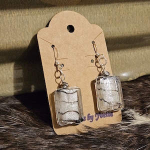 May include: A pair of silver dangle earrings with rectangular glass charms featuring a silver and white striped pattern. The earrings are displayed on a brown card with the text "by Trisha".