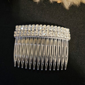 May include: A clear plastic hair comb with two rows of sparkling rhinestones across the top. The comb has multiple teeth and is set against a black background. This hair accessory is suitable for weddings or special occasions.