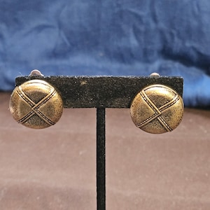 Puede incluir: Un par de pendientes de clip de tono dorado con un diseño circular texturizado. Los pendientes tienen un patrón de cruz en la parte delantera.