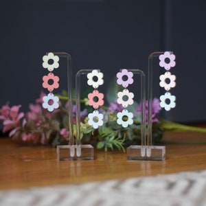 May include: Three pairs of dangle earrings with three flower charms each. The flowers are in pastel colors: light blue, light pink, and light yellow. The earrings are displayed on a clear acrylic stand.