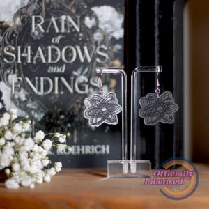 Puede incluir: Pendientes colgantes con un diseño abstracto. Cada pendiente presenta una forma circular transparente con un patrón negro garabateado. Los pendientes se exhiben en soportes transparentes. El fondo incluye un libro titulado "Rain of Shadows and Endings".