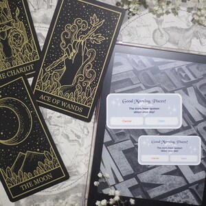 May include: Black and gold tarot cards with celestial illustrations, including "The Chariot", "Ace of Wands", and "The Moon". A tablet displays a message: "Good Morning, Pisces!" with "Cancel" and "Open" buttons. The cards are on a map.