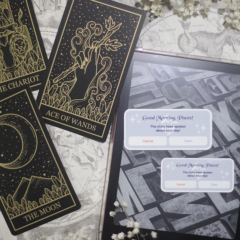 May include: Black and gold tarot cards with celestial illustrations, including "The Chariot", "Ace of Wands", and "The Moon". A tablet displays a message: "Good Morning, Pisces!" with "Cancel" and "Open" buttons. The cards are on a map.