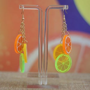 Puede incluir: Pendientes de cadena dorada con rodajas de cítricos coloridas. Los pendientes presentan rodajas de naranja, amarillo y verde, creando un accesorio lúdico y veraniego. Los pendientes se exhiben en un soporte transparente.