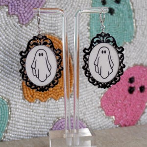 Op de afbeelding: Een paar zwarte en witte geestoorbellen met een decoratief frame. De oorbellen hangen aan een transparante standaard.