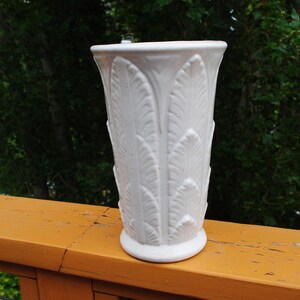 May include: A white ceramic vase with a flared top and a round base. The vase features a repeating embossed feather design. The vase is approximately 10 inches tall and sits on a wooden surface with a green background.