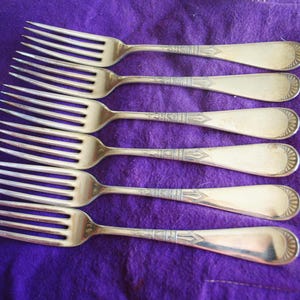 May include: Six antique silver forks arranged on a purple cloth. The forks have a decorative design on the handles and a classic, elegant shape. The tines are long and slender. The forks are likely for a formal dining setting.
