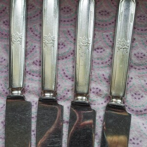 May include: Four silver-toned metal dinner knives arranged side-by-side. Each knife features a flat, rectangular handle with a vertical grooved pattern and a small, embossed floral motif near the center. The blades are smooth and reflective.