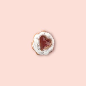 May include: A single heart-shaped cookie with a red jelly center, surrounded by white frosting. The cookie has a golden-brown edge and sits on a light pink background. A sweet treat.
