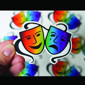 May include: A sticker featuring two theater masks: a smiling mask in rainbow colors and a sad mask in blue and green. The masks are connected by a black swirl design. Other masks are visible in the background.