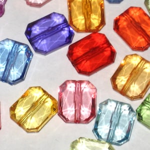 May include: Assortment of colorful faceted glass beads in various shades of purple, pink, blue, red, yellow, and orange. The beads are rectangular in shape and have a hole in the center for stringing.