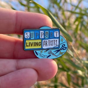 May include: A blue and yellow enamel pin with the text "Support Living Artists" and a skull and crossbones symbol. The pin also features a variety of artistic tools, including a paintbrush, a pencil, a needle, and a pair of scissors.