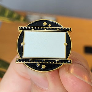 May include: A gold-toned metal pin with a black and white design. The pin features a circular frame with a black border and a white rectangular center. The design includes gold accents and a ruler-like detail along the top and bottom edges.