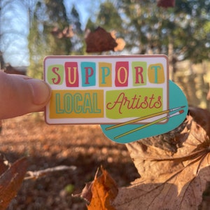May include: A sticker with the words "Support Local Artists" in colorful block letters. The sticker has a blue background with two paintbrushes crossed over each other.