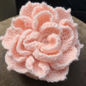 May include: A handmade, light pink crochet flower with multiple layers of petals. The petals are tightly wound, creating a full, textured appearance. The craftwork showcases a soft, delicate aesthetic, perfect for home decor or as a unique gift.