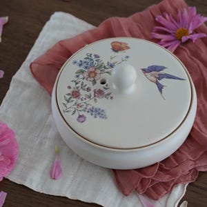 May include: A white ceramic tea caddy with a lid featuring a floral and bird design. The lid has a small white knob and a butterfly illustration. The floral design includes pink, blue, and purple flowers. The caddy is on a pink cloth.