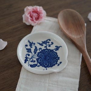 May include: A white ceramic spoon rest with a blue floral design. The spoon rest is round with a small indentation for the spoon handle. A wooden spoon and a pink rose are also in the image.