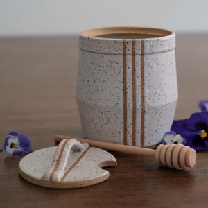 May include: White ceramic honey pot with a brown lid and a wooden honey dipper. The pot has two brown stripes around it.