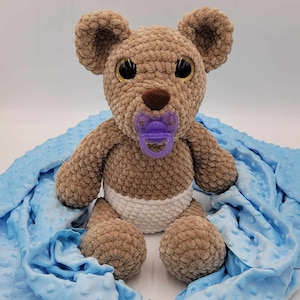 May include: A brown crocheted teddy bear with large eyes and a purple pacifier. The bear is sitting on a light blue blanket.