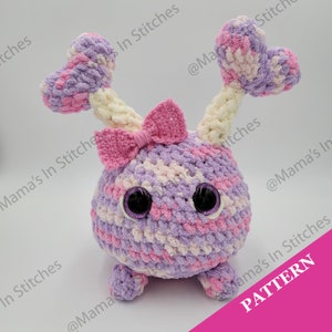 May include: A crocheted plush toy with a purple, pink, and white color scheme. The toy has a heart-shaped head with two antennas, a pink bow, and large black eyes. The toy has four legs and a round body. The toy is sitting on a white surface. The text "PATTERN" is written in pink letters on a pink background.