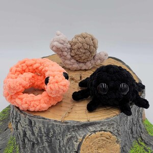 May include: Three crocheted stuffed animals on a tree stump. A pink and orange worm, a black spider, and a light brown snail.