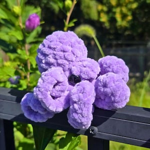 Könnte beinhalten: Ein handgefertigtes, flauschiges lila Oktopus-Spielzeug mit rundem Kopf, acht Tentakeln und einem schwarzen Perlenauge. Das weiche, flauschige Spielzeug sitzt auf einem schwarzen Metallgeländer, mit grünem Laub im Hintergrund.