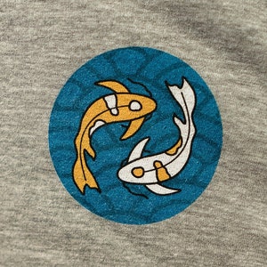May include: Close-up of a gray t-shirt featuring a circular emblem with two koi fish. One fish is orange, the other is white with orange accents, set against a teal background with a wave pattern. The shirt is made of a soft, textured fabric.