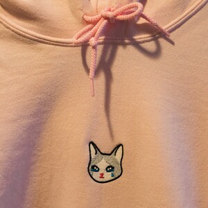 Op de afbeelding: Een lichtroze hoodie met een capuchon met trekkoord en een kangoeroezak. De hoodie heeft een geborduurde patch van een huilende kattenkop op de borst. De kat heeft blauwe tranen en een grijs en wit gezicht.