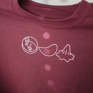 May include: A maroon sweatshirt with a crew neck and white graphic. The graphic features three food-related line drawings: a chicken in a hat, a potato chip, and a blob.