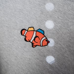 Peut inclure: Un t-shirt gris avec un motif de poisson-clown brodé en couleur. Le poisson est orange avec des rayures blanches et a des nageoires bleues et jaunes.