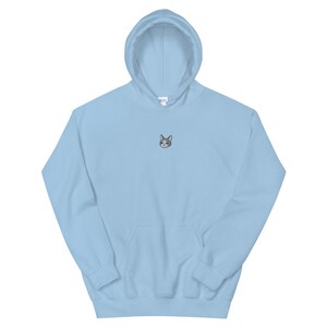 Op de afbeelding: Een lichtblauwe hoodie met een voorzak en een capuchon. De hoodie heeft een kleine geborduurde kattenkop op de borst. De kattenkop is grijs en wit met blauwe ogen. De hoodie is gemaakt van een zacht materiaal.