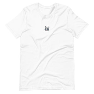 Op de afbeelding: Wit t-shirt met een kleine geborduurde kattenkop. De kat heeft grijs en wit haar, blauwe ogen en een roze neus. Het t-shirt is gemaakt van een zachte, lichte stof.