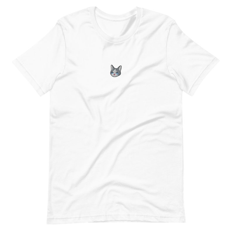 Op de afbeelding: Wit t-shirt met een kleine geborduurde kattenkop. De kat heeft grijs en wit haar, blauwe ogen en een roze neus. Het t-shirt is gemaakt van een zachte, lichte stof.