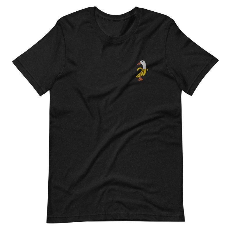 Może przedstawiać: Czarny t-shirt z małym haftowanym wzorem ptaka z białą głową i ż&oacute;łtym bananowym korpusem. Ptak ma pomarańczowe stopy i jest zwr&oacute;cony w prawo. T-shirt jest wykonany z miękkiego materiału.