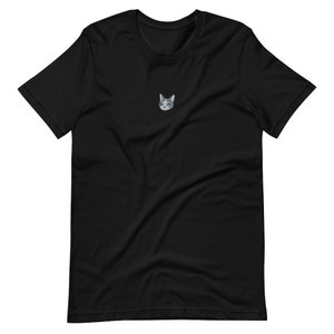 Op de afbeelding: Zwart t-shirt met een kleine geborduurde kattenkop. De kat heeft grijs en wit haar, blauwe ogen en een kleine traan. Het t-shirt is gemaakt van een zacht materiaal en heeft een klassieke ronde hals.
