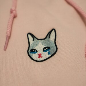 Op de afbeelding: Een lichtroze hoodie met een trekkoord en een kattengezicht geborduurd op de voorkant. De kat heeft een grijs en wit gezicht met blauwe ogen en een rode mond en huilt een traan.