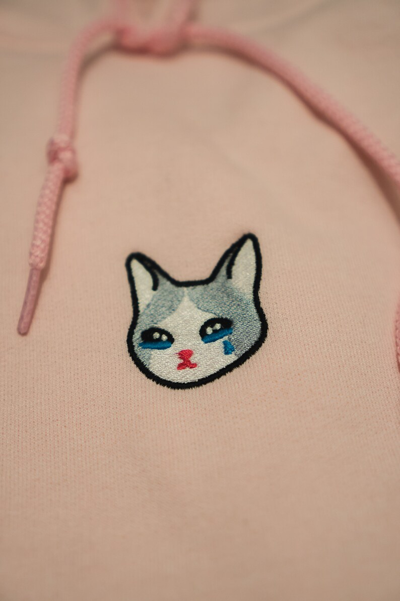 Op de afbeelding: Een lichtroze hoodie met een trekkoord en een kattengezicht geborduurd op de voorkant. De kat heeft een grijs en wit gezicht met blauwe ogen en een rode mond en huilt een traan.