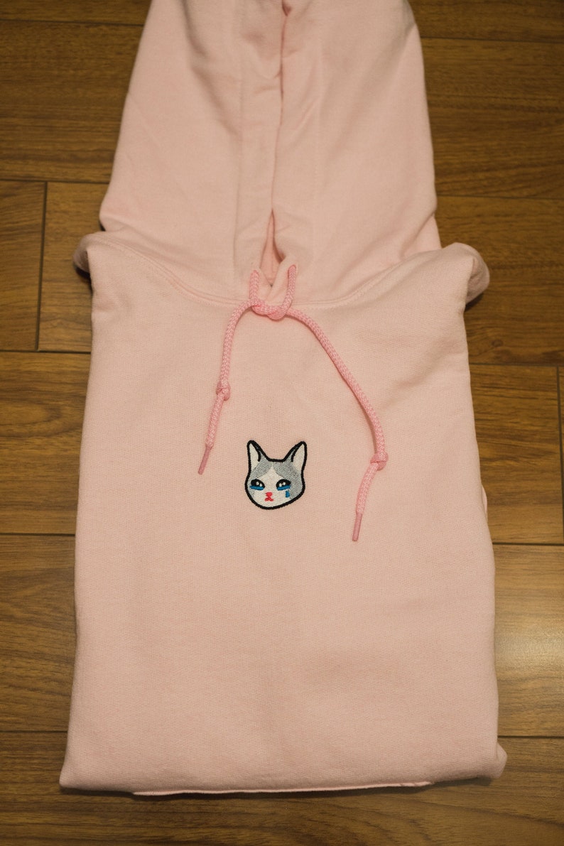 Op de afbeelding: Een lichtroze hoodie met een capuchon met trekkoord. De voorkant heeft een geborduurd kattengezicht met blauwe tranen. De hoodie is op een houten oppervlak gevouwen. Het trekkoord en de details van het kattengezicht zijn roze.