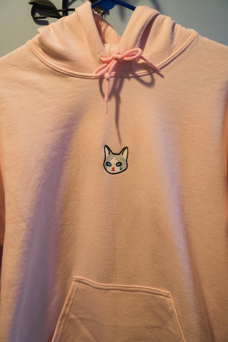 Op de afbeelding: Een lichtroze hoodie met een capuchon met trekkoord en een kangoeroezak. De hoodie heeft een geborduurde patch van een huilende kattenkop op de borst. De kat heeft blauwe tranen en een grijs en wit gezicht.