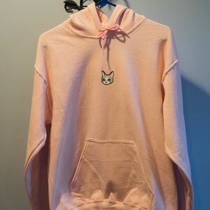 Op de afbeelding: Een lichtroze hoodie met een voorzak en een capuchon met trekkoord. De hoodie heeft een geborduurd kattengezicht op de borst. Hij hangt aan een blauwe hanger tegen een blauwe muur.