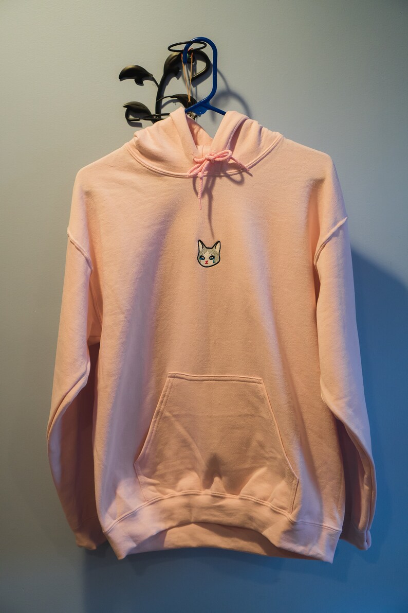 Op de afbeelding: Een lichtroze hoodie met een voorzak en een capuchon met trekkoord. De hoodie heeft een geborduurd kattengezicht op de borst. Hij hangt aan een blauwe hanger tegen een blauwe muur.
